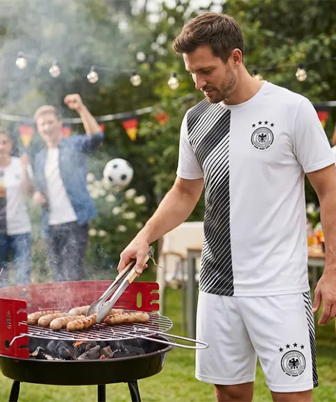 Ein Mann in Sportkleidung grillt Würstchen auf einem Holzkohlegrill in einem Garten, im Hintergrund Menschen und Dekorationen zum Thema Fußball.